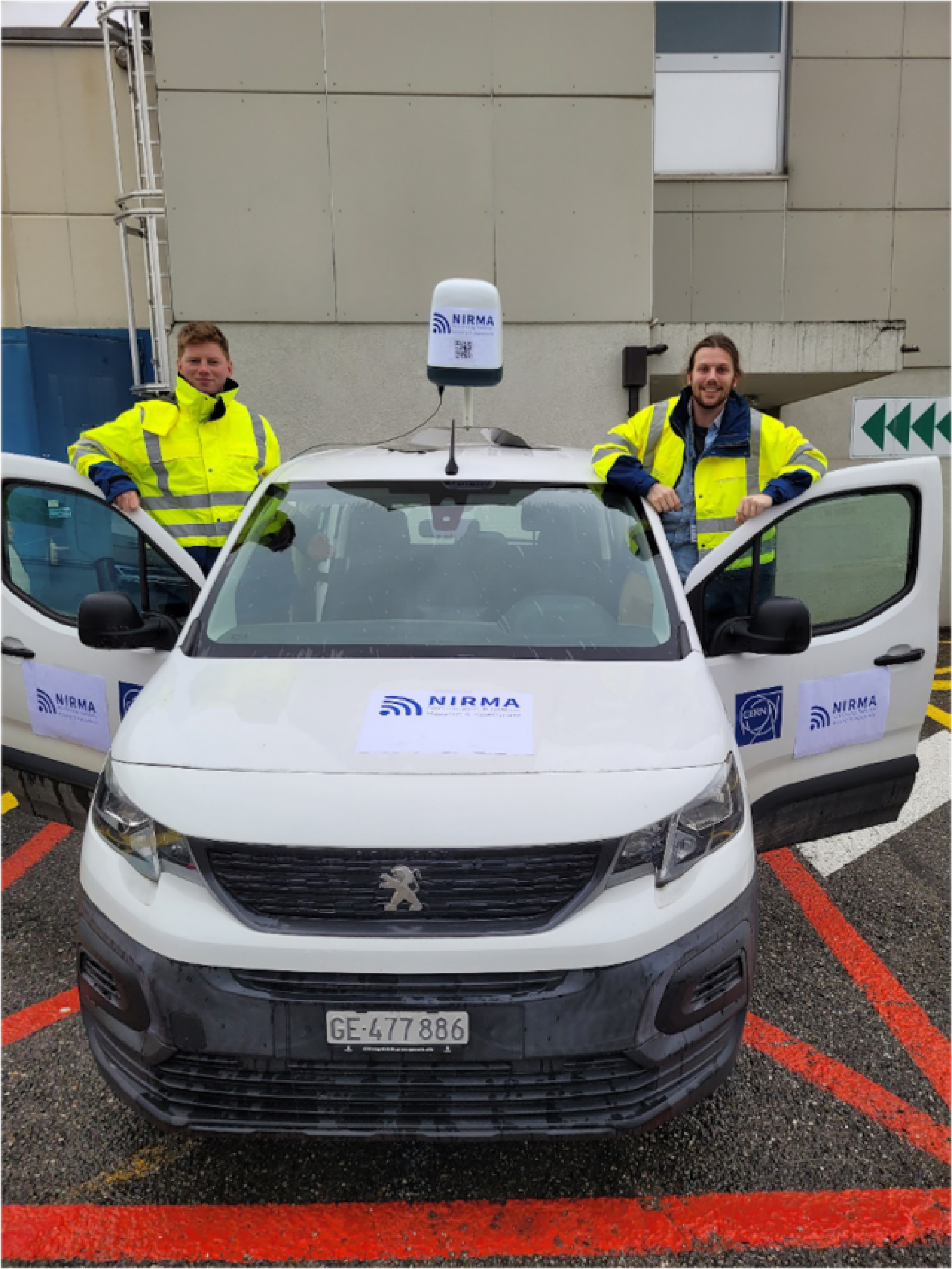 The students, in high visibility, smiling whilst standing behind the doors of a small white van. Upon which is mounted a small white cube, the NIRMA sensor. The vehicle displays the NIRMA logo and QR codes for more info.